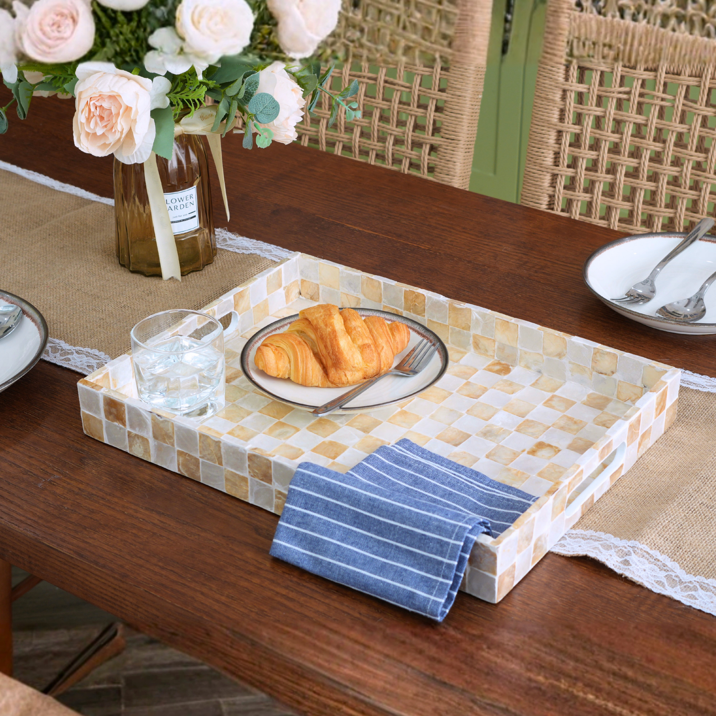 A rectangular serving tray with a mosaic tile pattern in beige, cream, and light brown sits on a wooden dining table. The tray is styled with a blue-and-white striped napkin, a plate with a croissant, a glass of water, and a vase of flowers in the background.