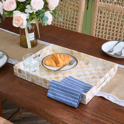 A rectangular serving tray with a mosaic tile pattern in beige, cream, and light brown sits on a wooden dining table. The tray is styled with a blue-and-white striped napkin, a plate with a croissant, a glass of water, and a vase of flowers in the background.