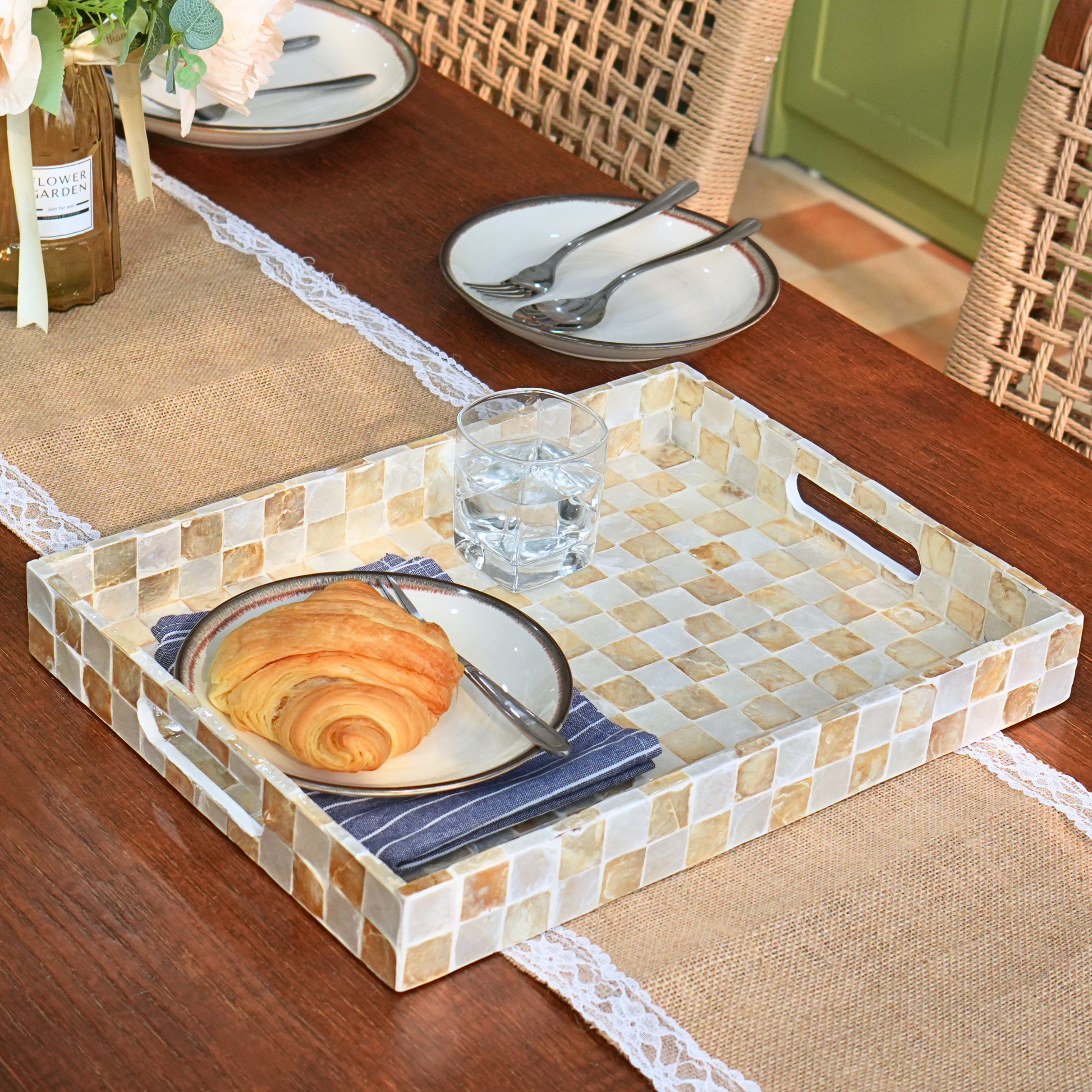 A rectangular serving tray with a mosaic tile pattern in beige, cream, and light brown, styled on a wooden table with plates, croissants, a glass of water, and a blue napkin.