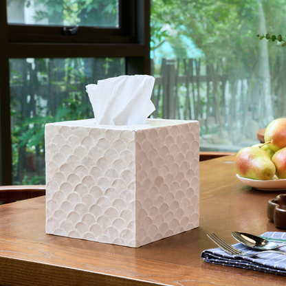 A square white tissue box cover with a textured, raised scallop pattern sits on a wooden table in a kitchen or dining setting, with tissue paper pulled from the top. A plate of apples and folded napkins are visible in the background.