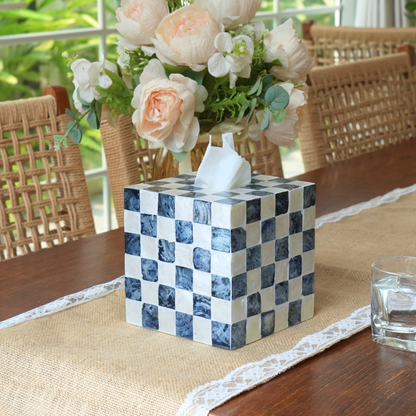 A kleebex tissues cube boxes cover with a blue and white checkerboard pattern sits on a wooden table runner, styled with a vase of peach and white flowers in the background.
