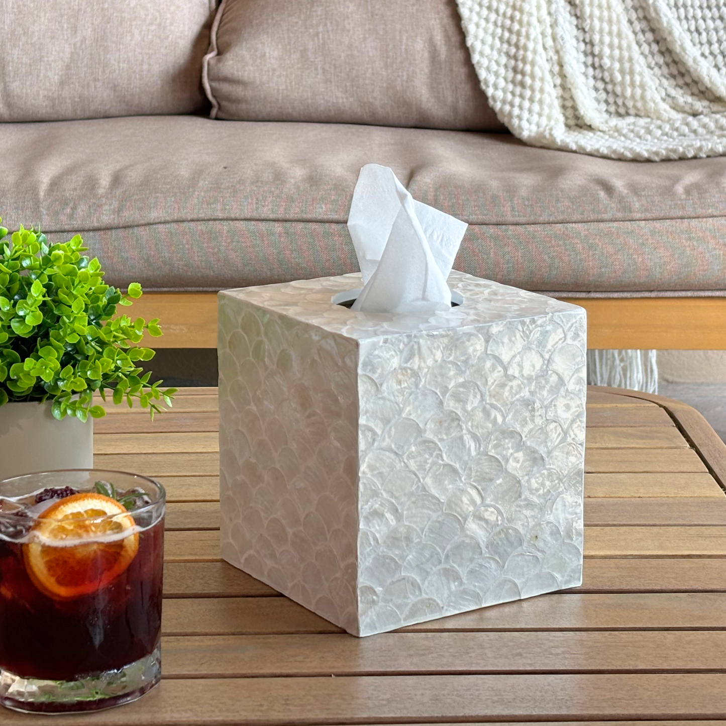 A kleenex tissues cube boxes cover with a capiz shells surface and a white tissue protruding from the top, placed on a wooden table in a living room setting with a sofa, plant, and drink in the background.