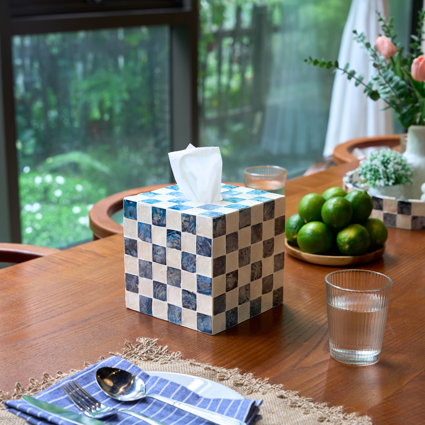 A kleenex cube cover with navy blue and white checkered pattern sits on a wooden dining table. The box has a white tissue protruding from the top, and the scene includes a bowl of limes, a glass of water, and a blue-striped napkin in a real indoor environment.