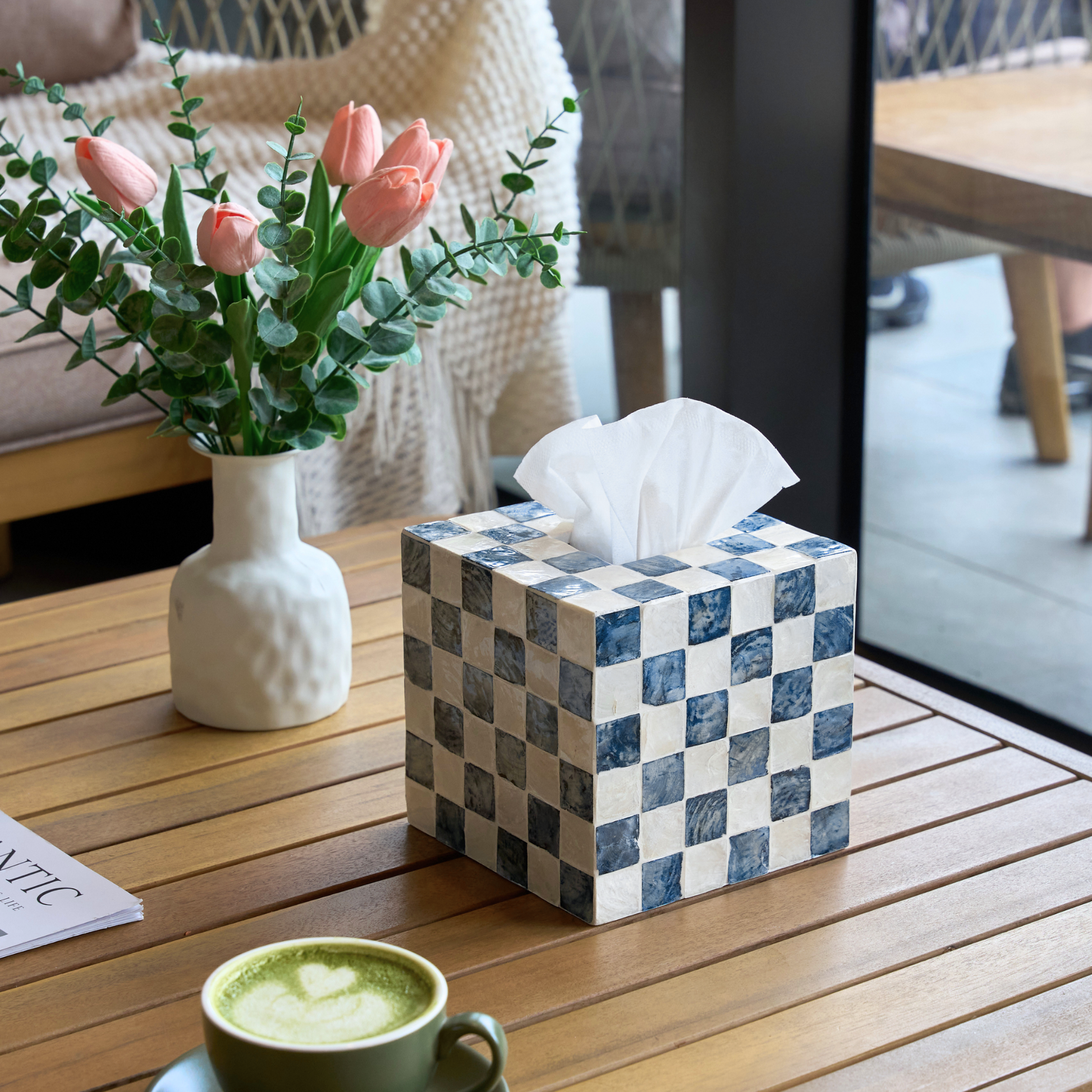 A square tissue cover box with blue and white checkered tiles sits on a wooden table, with a white tissue protruding from the top. A white vase with pink tulips and a green cup are nearby, and the background shows a cozy indoor setting with a window and furniture.