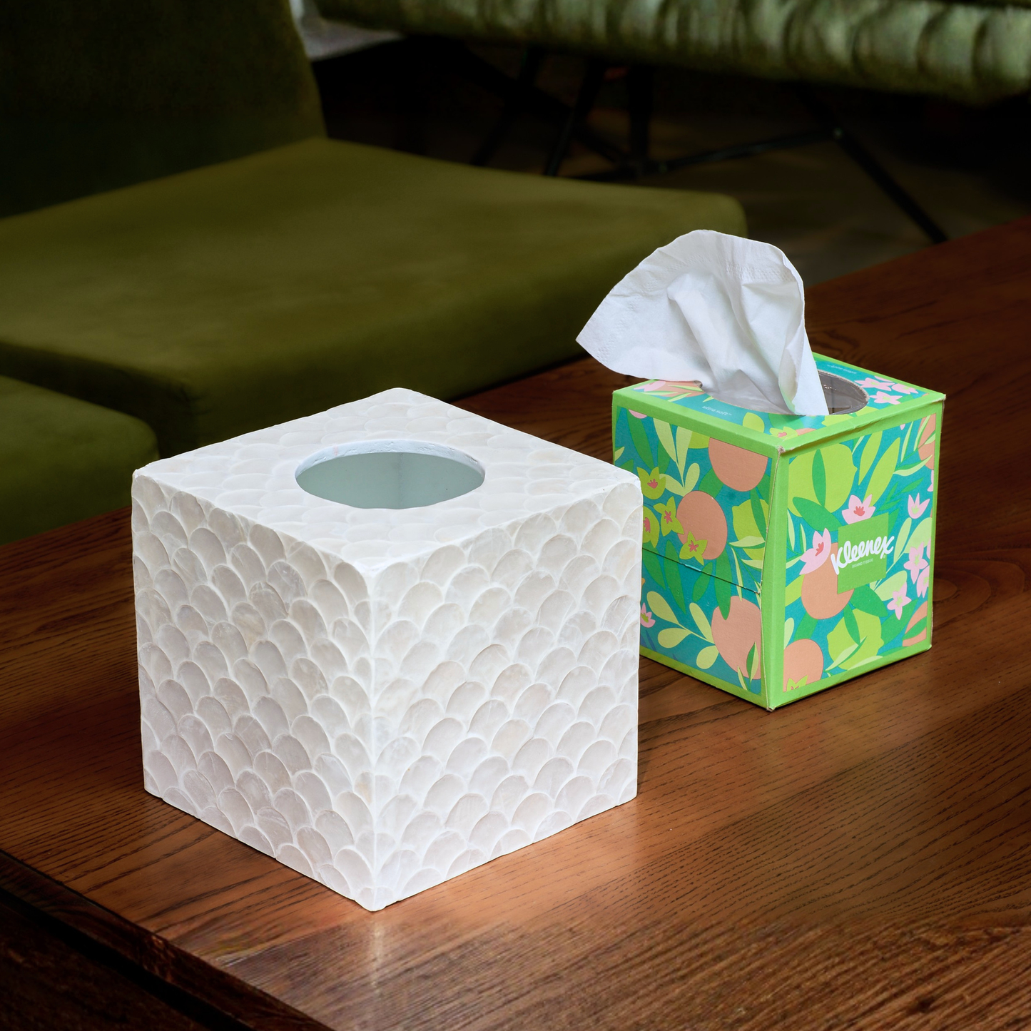 A white square tissue box cover coastal  sits on a wooden table next to a green Kleenex tissue box with a white tissue protruding, with an olive green sofa in the background.