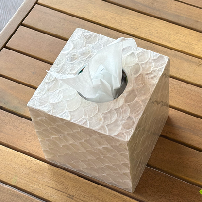 A square capiz tissue box cover with a raised scalloped texture, shown with a white tissue protruding from the top, placed on a brown wooden slatted surface.