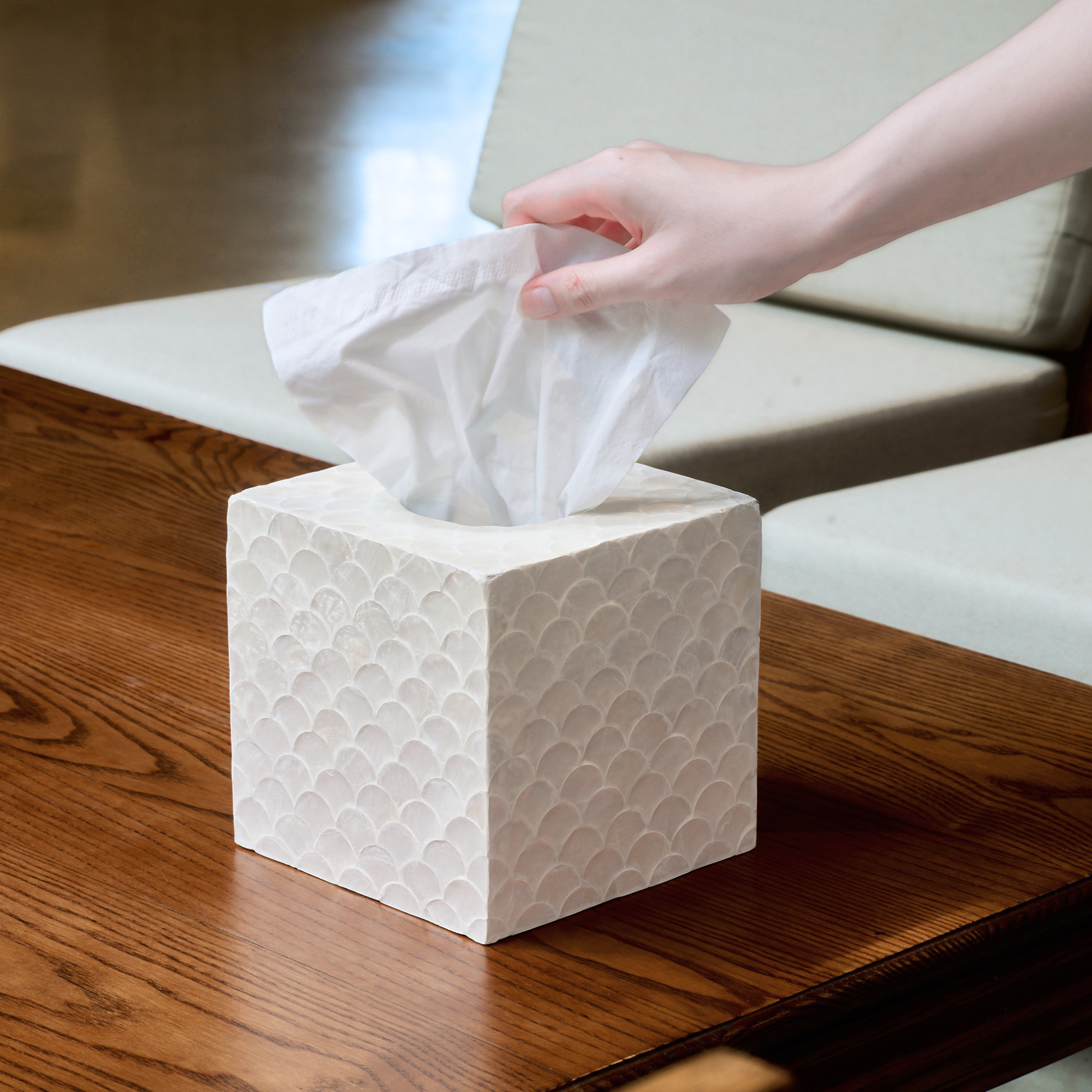 A cute tissue box cover in white color with a textured pattern sits on a wooden table, with a hand pulling a white tissue from the top.