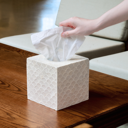 A cute tissue box cover in white color with a textured pattern sits on a wooden table, with a hand pulling a white tissue from the top.