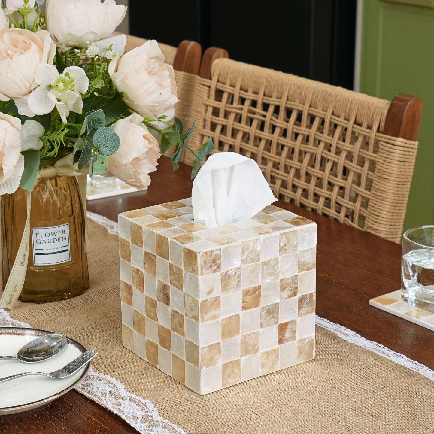 A square tissue box cover made of beige and ivory checkered capiz shells tiles sits on a dining table, with a white tissue protruding from the top. A flower vase, woven chairs, placemat, and glass are visible around it in a real indoor setting.