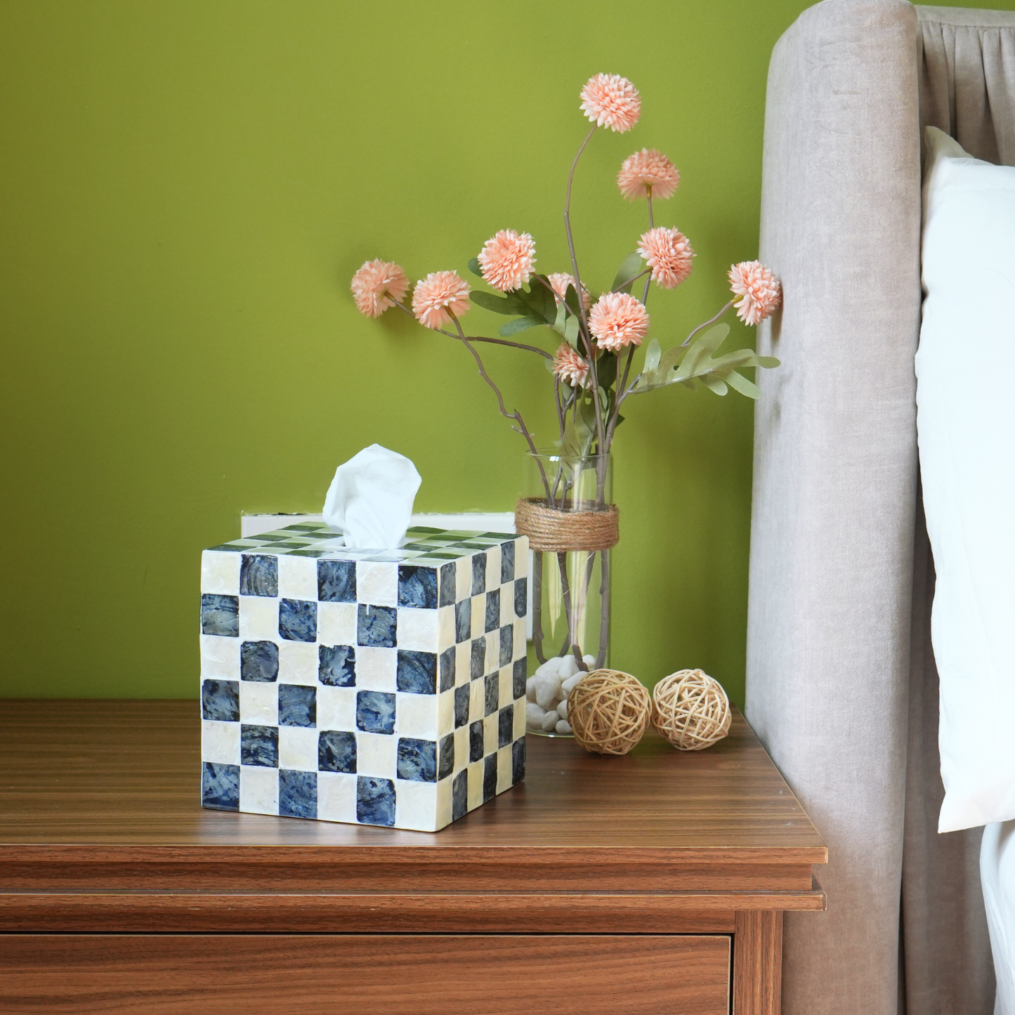 A checkered tissue box cover in blue and white sits on a wooden nightstand beside a glass vase with pink flowers, against an olive green wall and next to a bed.
