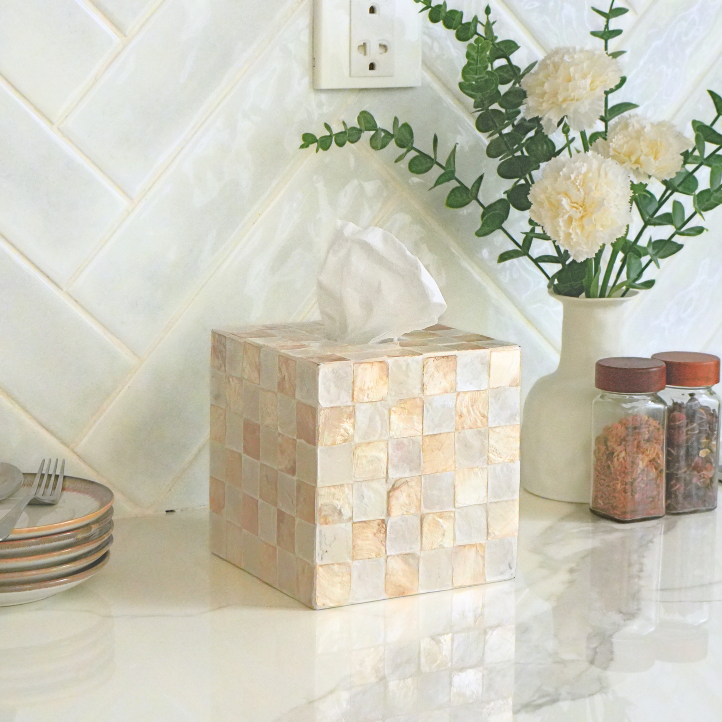 A square tissue box cover made of small beige and cream mosaic tiles sits on a white countertop in a kitchen setting, with a vase of flowers and spice jars behind it.