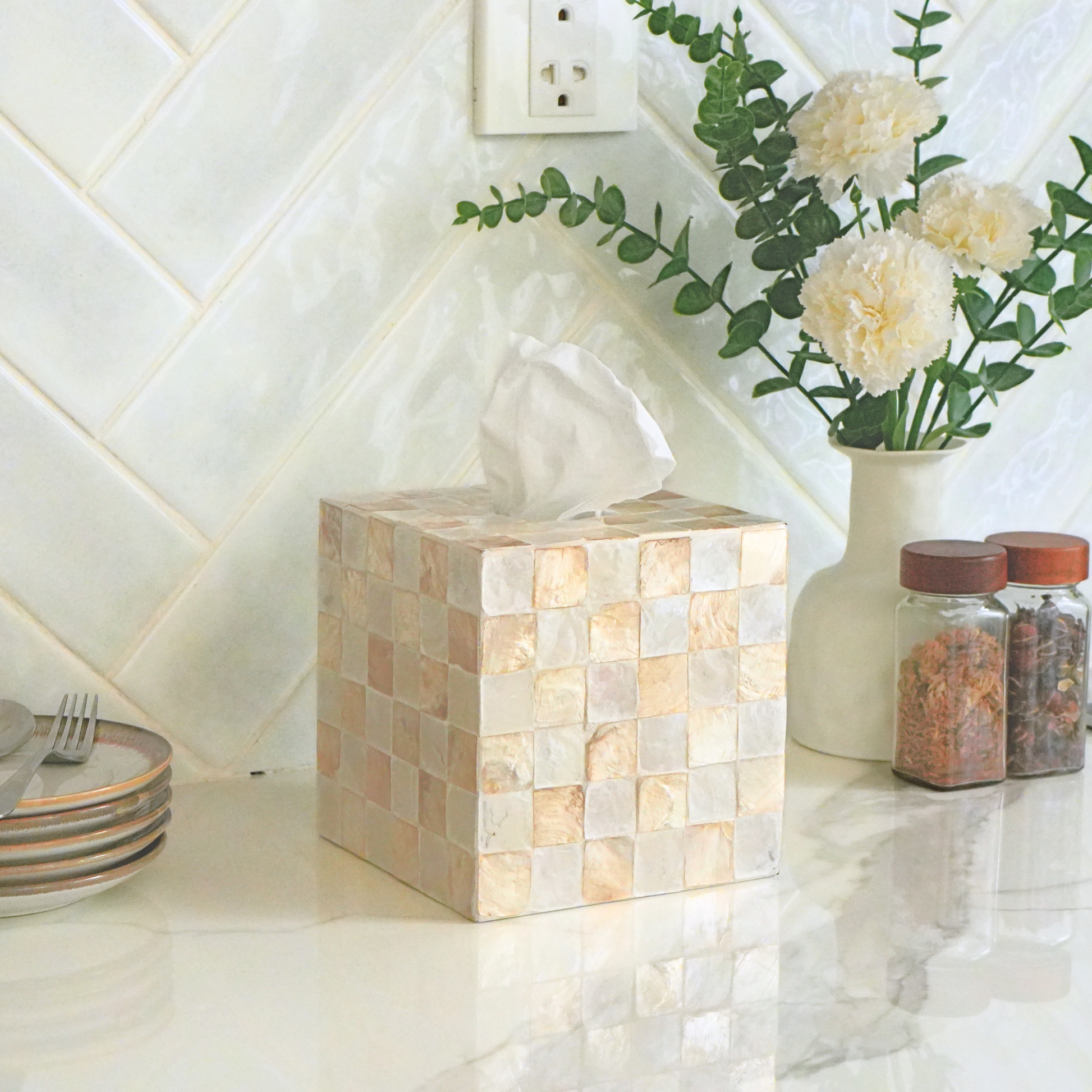 A square tissue box cover made of small beige and cream mosaic tiles sits on a white countertop in a kitchen setting, with a vase of flowers and spice jars behind it.