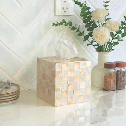 A square tissue box cover made of small beige and cream mosaic tiles sits on a white countertop in a kitchen setting, with a vase of flowers and spice jars behind it.