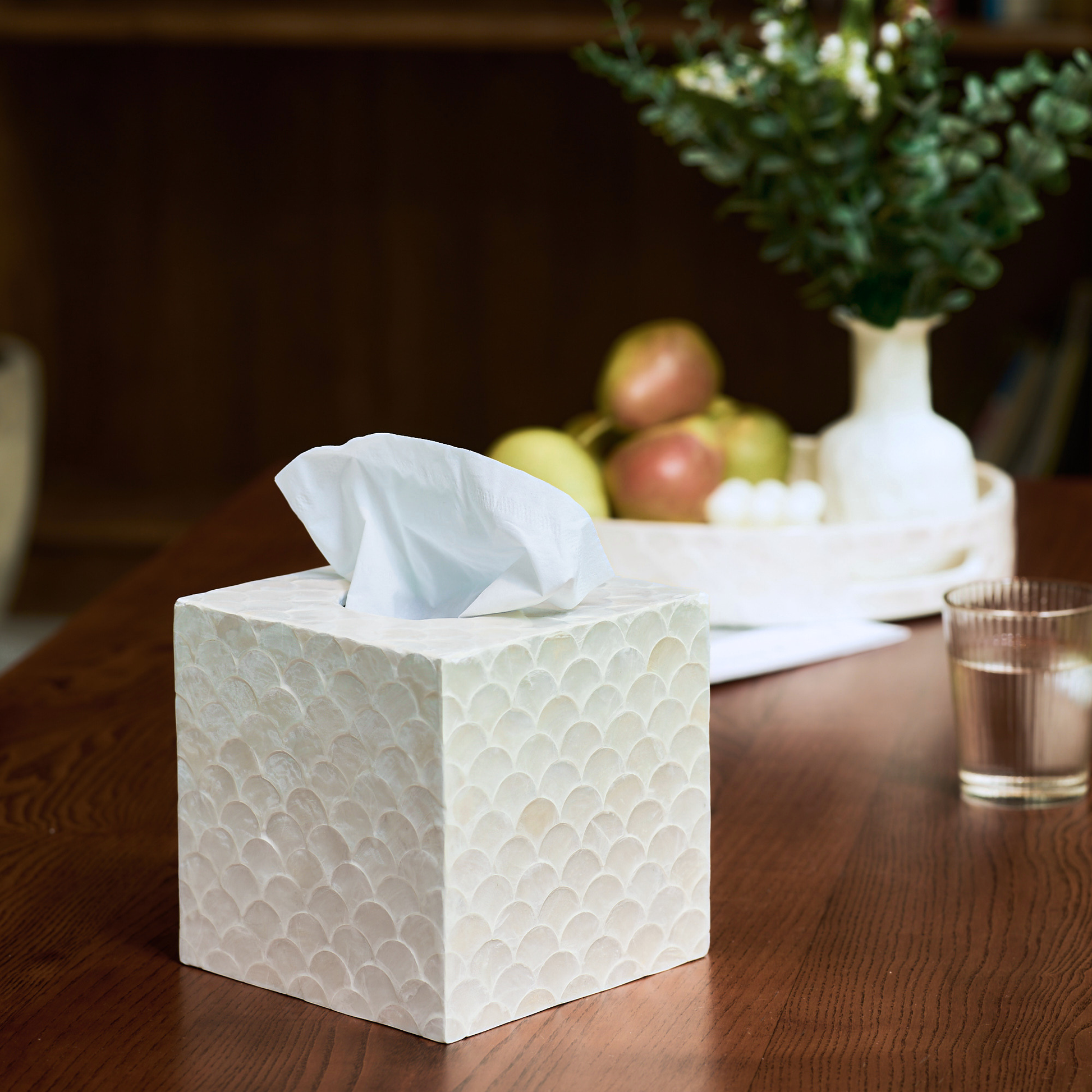 A square white tissue box cover with a textured fish scale pattern sits on a wooden table, with a white tissue protruding from the top. In the softly blurred background, a white vase with greenery and fruit on a tray are visible.