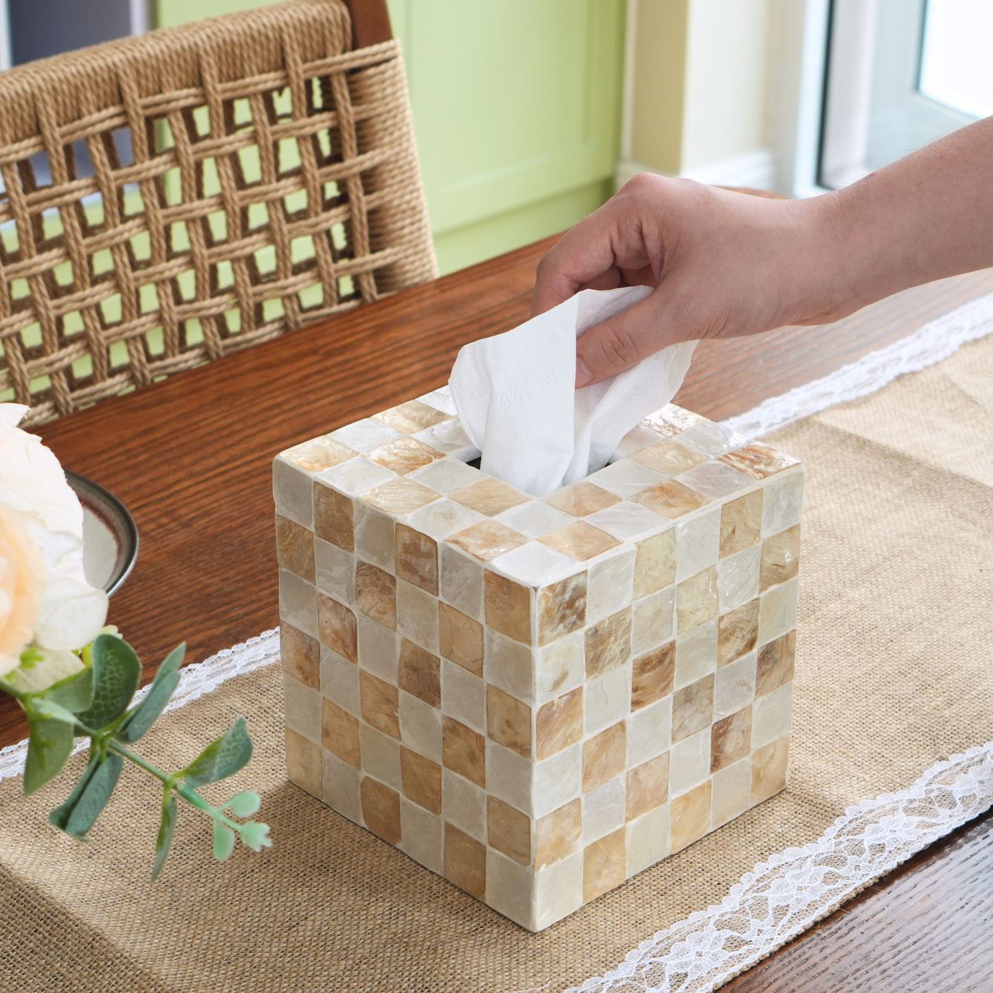 A boho tissue box cover made of small beige and cream mosaic tiles, shown on a table with a hand pulling a white tissue out of the top. A woven chair and a flower are visible in the background.