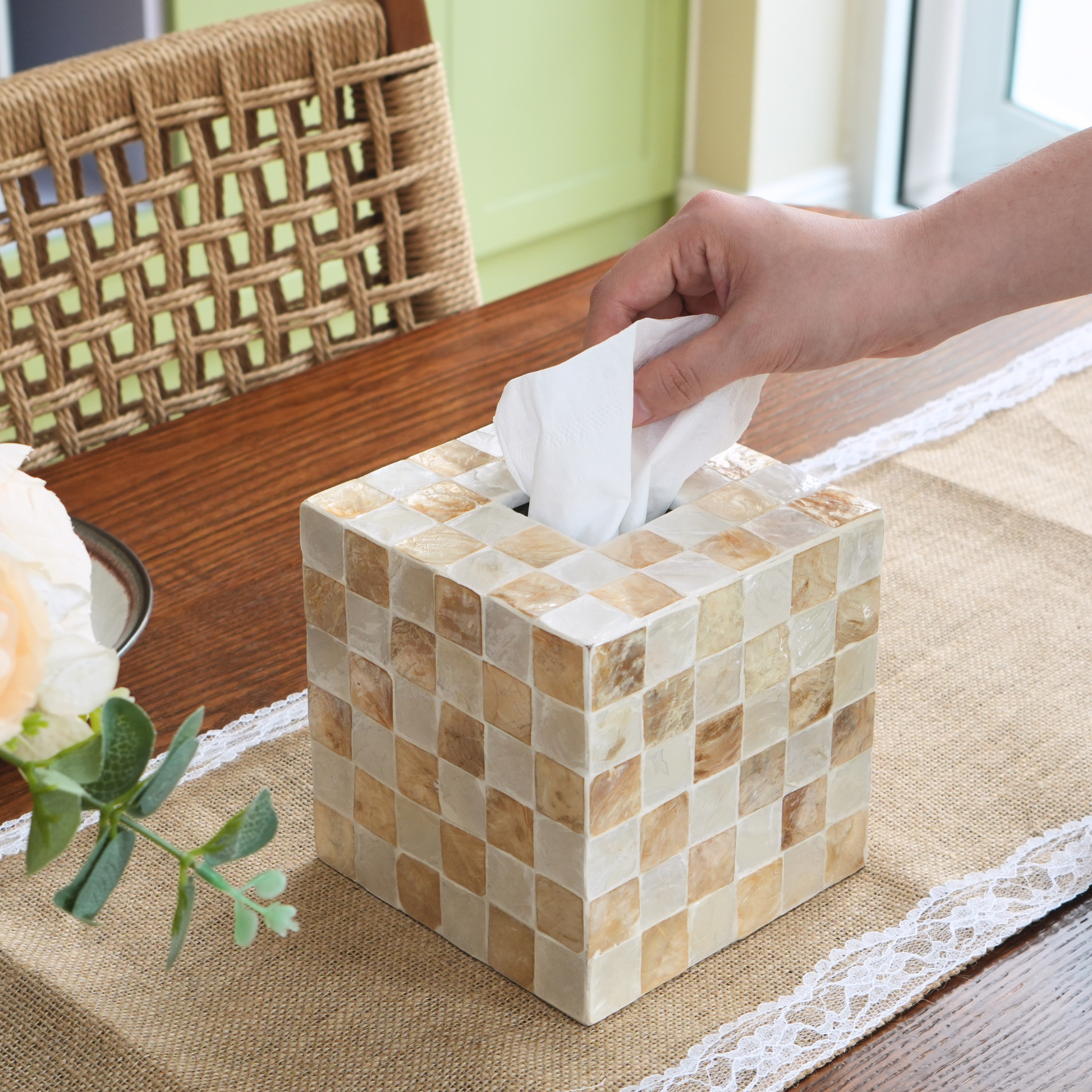 A boho tissue box cover made of small beige and cream mosaic tiles, shown on a table with a hand pulling a white tissue out of the top. A woven chair and a flower are visible in the background.