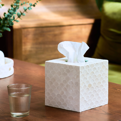 A white textured tissue box holder with a tissue protruding from the top, placed on a wooden table next to a glass of water in a softly lit indoor setting.