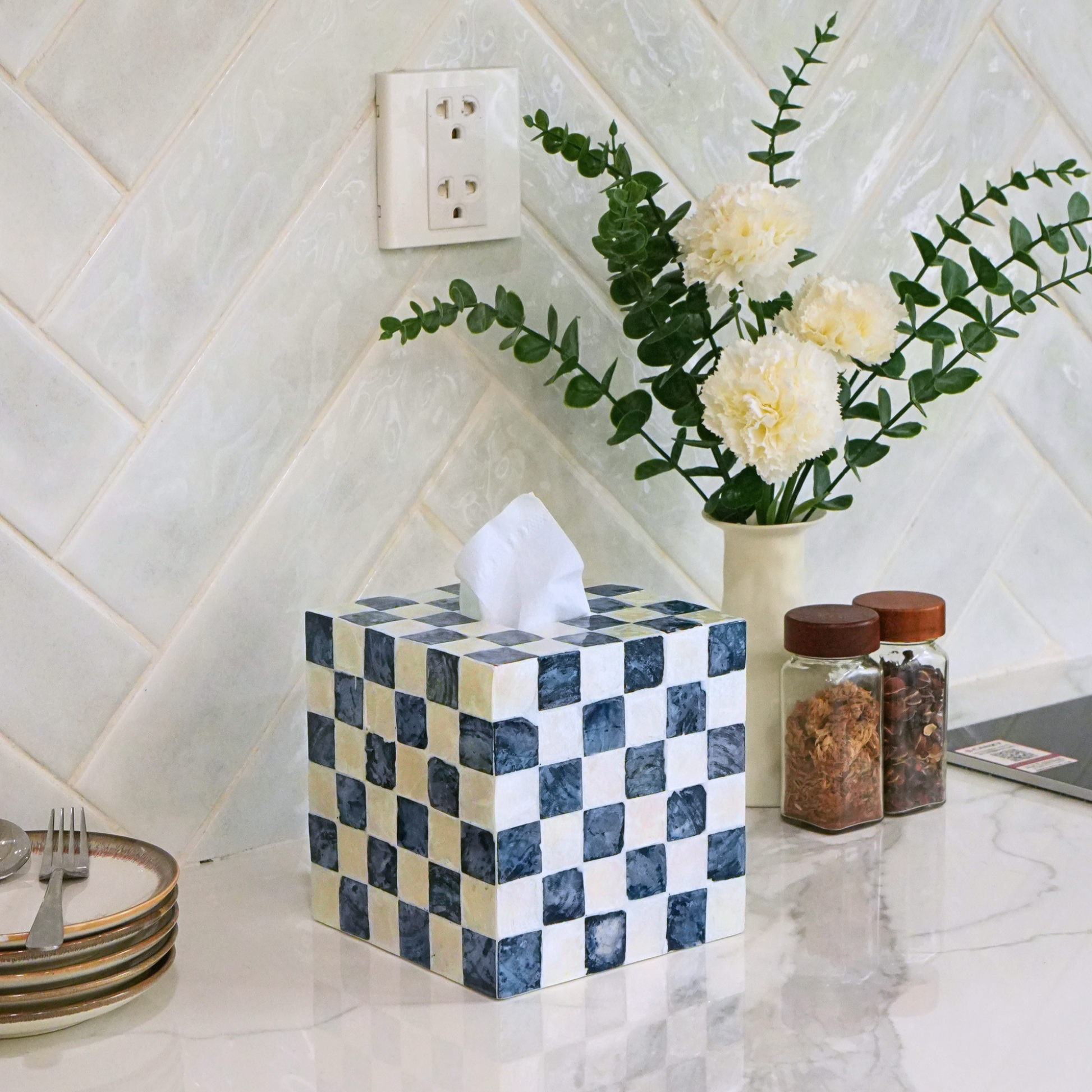 A square tissue box cover with a blue and cream checkered pattern sits on a marble countertop. A white tissue is pulled from the top, next to a vase of white flowers and two spice jars, against a white tiled wall.