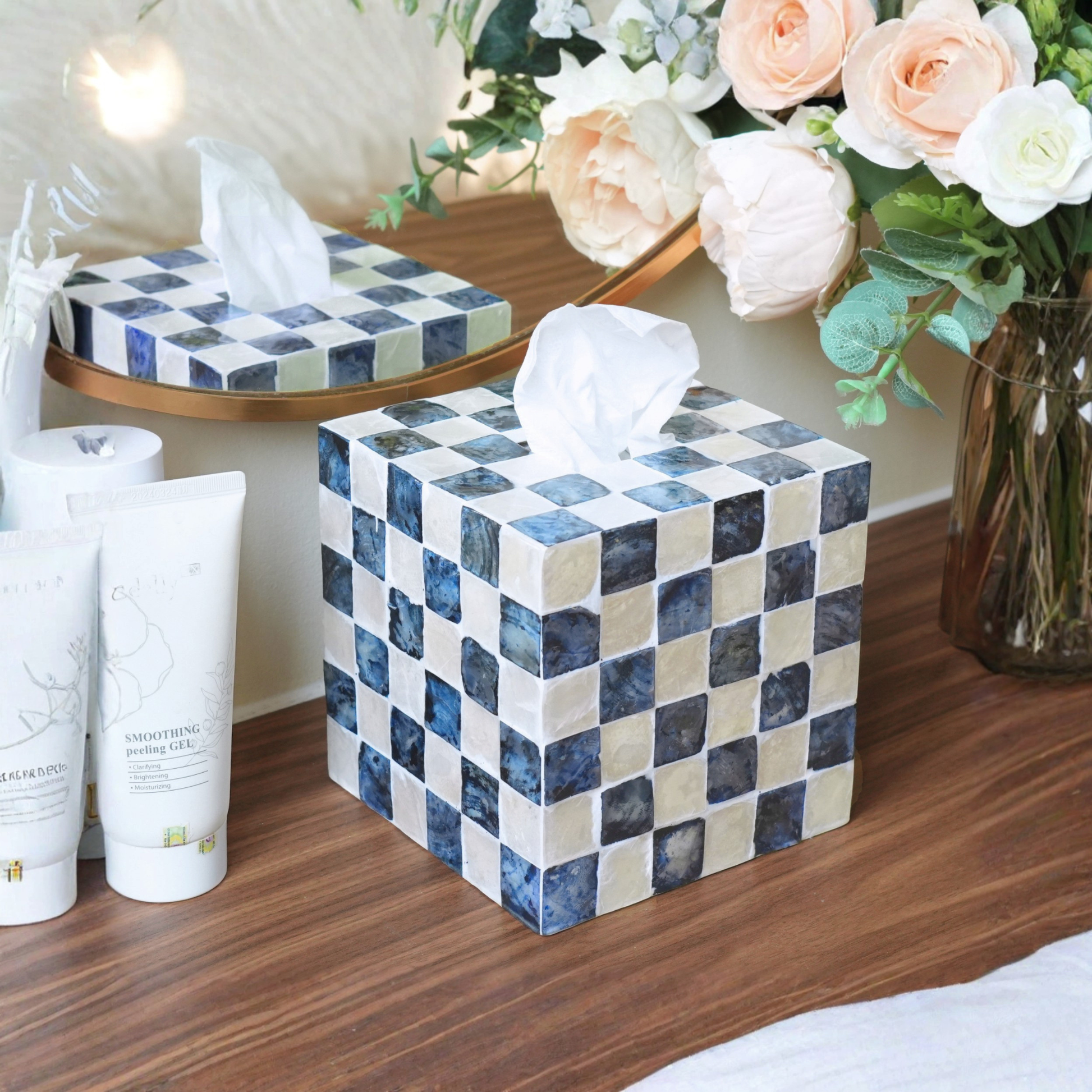 A square tissue box cover with a blue and beige capiz shell checkerboard pattern sits on a wooden table next to skincare tubes and a mirror, with a vase of flowers in the background.