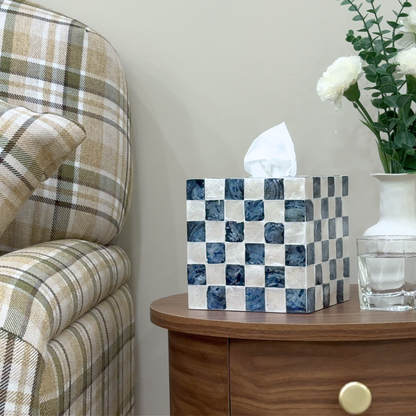 Navy Blue tissue box cover with checkered design on a wooden table next to a plaid chair and vase with flowers.