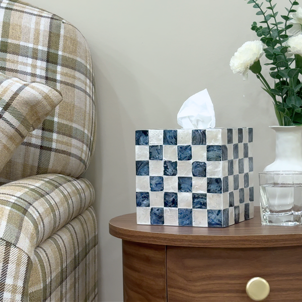 Navy Blue tissue box cover with checkered design on a wooden table next to a plaid chair and vase with flowers.