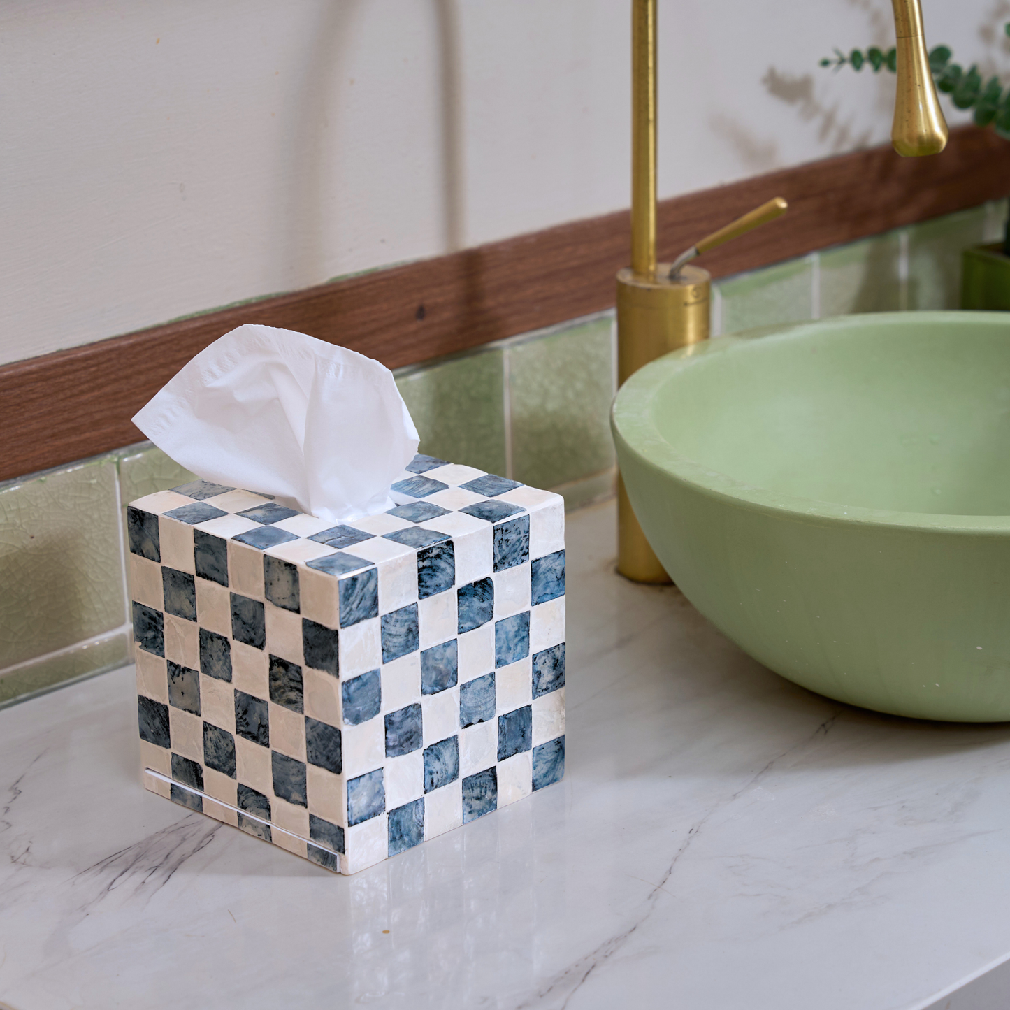 A square bathroom tissue holder made of blue and white checkered capiz shells tiles sits on a white marble bathroom counter next to a light green vessel sink and a gold faucet.