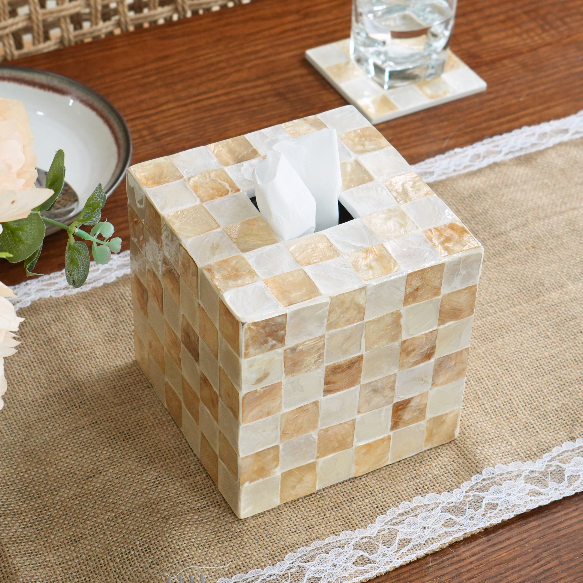 A square tissue box holder made of beige and cream checkered capiz shells tiles, shown holding white tissues on a burlap table runner with a glass and plate nearby.