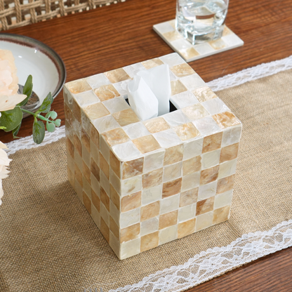 A square tissue box holder made of beige and cream checkered capiz shells tiles, shown holding white tissues on a burlap table runner with a glass and plate nearby.