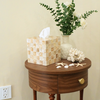 A mother of pearl tissue box cover made of beige and cream checkered shell tiles sits on a dark wood side table next to a vase of white flowers and starfish decorations.